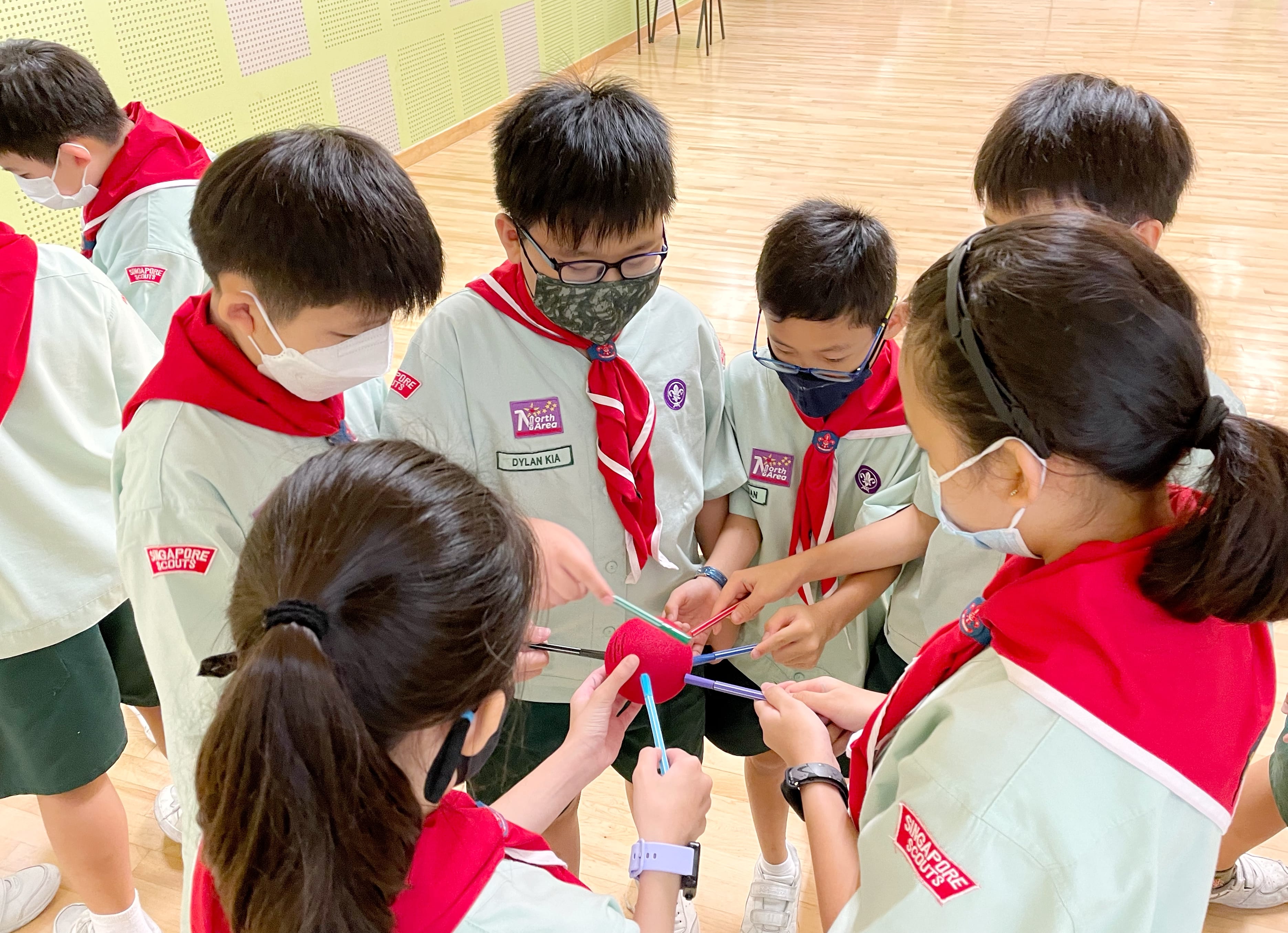 A group of masked Singapore Scouts children in uniform huddle together handling sticks and a red ball indoors.
